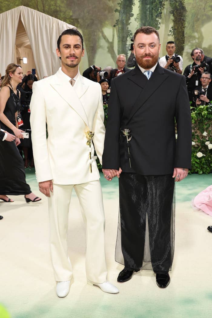 Sam Smith and Christian Cowan in formal wear, holding hands at an event, posing for photos and surrounded by attendees