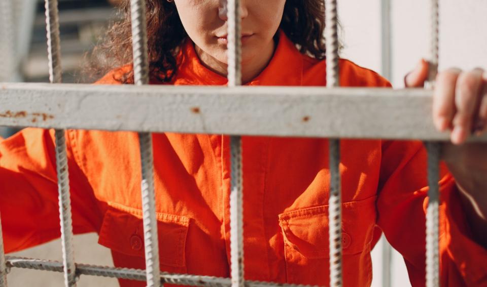 Person in orange outfit holding onto a metal gate with a serious expression