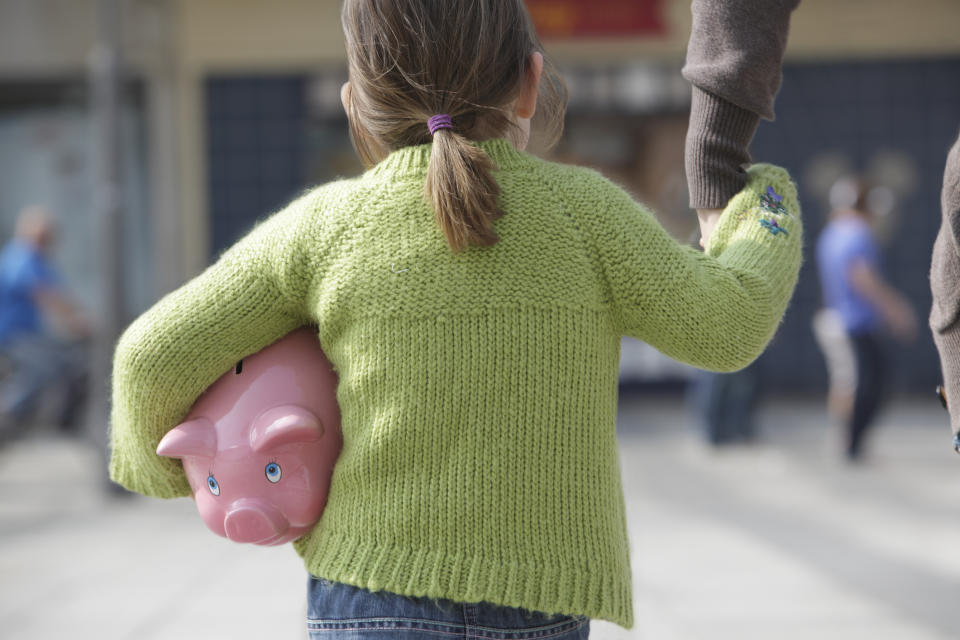 A child holding a pink piggy bank is seen from behind, wearing a knit sweater while walking hand-in-hand with an adult