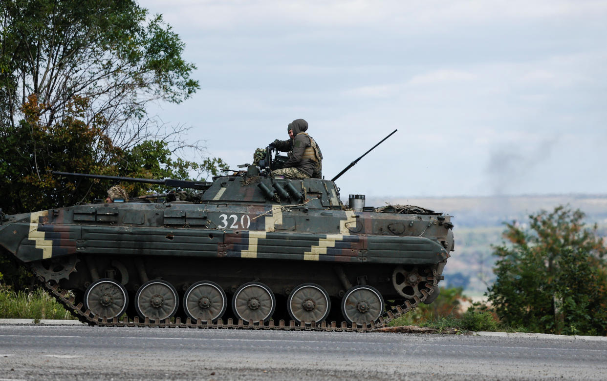 Two Ukrainian servicemen riding a BMP-2 infantry fighting vehicle along a road in the middle of agricultural land, with a blast in the distance.