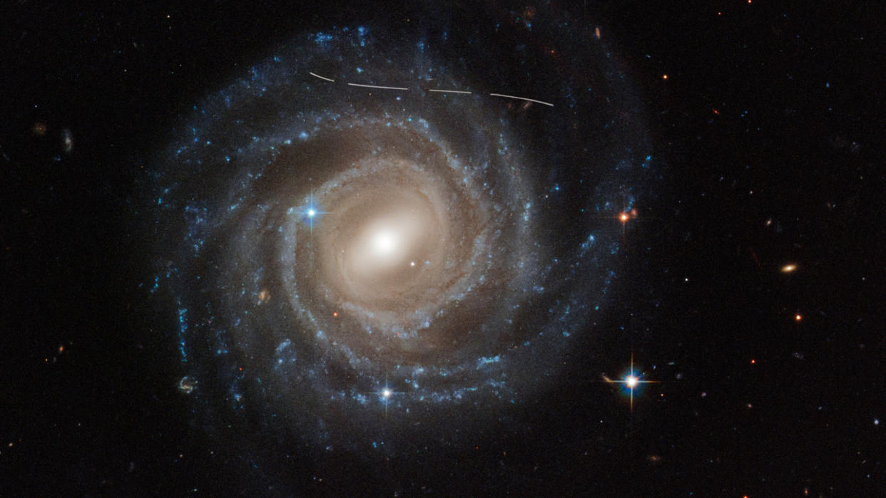  Hubble space telescope of a spiral galaxy in deep space, with a dotted line near its top indicating the trail of an asteroid in our own solar system. 