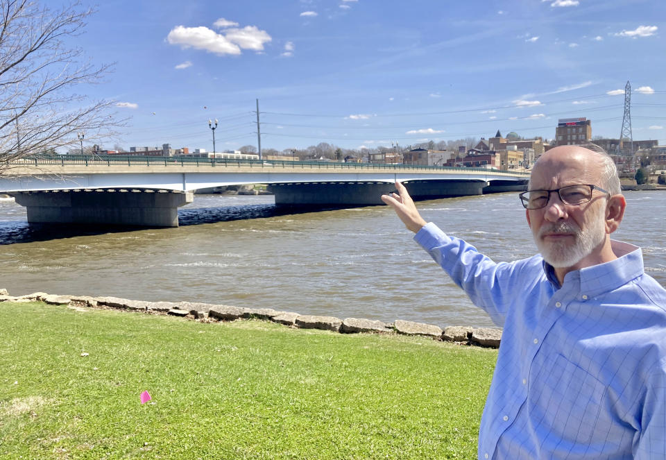 Tom Wadsworth gestures towards the Truesdell bridge while standing on the bank of the Rock River in Dixon, Ill., on Tuesday, April 11, 2023, near the site of the May 4, 1873, baptismal ceremony, when a large crowd gathered on the bridge before it toppled over, killing 46 and injuring 56 in the worst road-bridge disaster in American history. Wadsworth's great-grandmother, Gertie Wadsworth, was 3 1/2 years old and was in the arms of her grandmother, Christan Goble, the day the bridge collapsed. Goble died, but Gertie was plucked from the river downstream and revived. (AP Photo/John O'Connor)