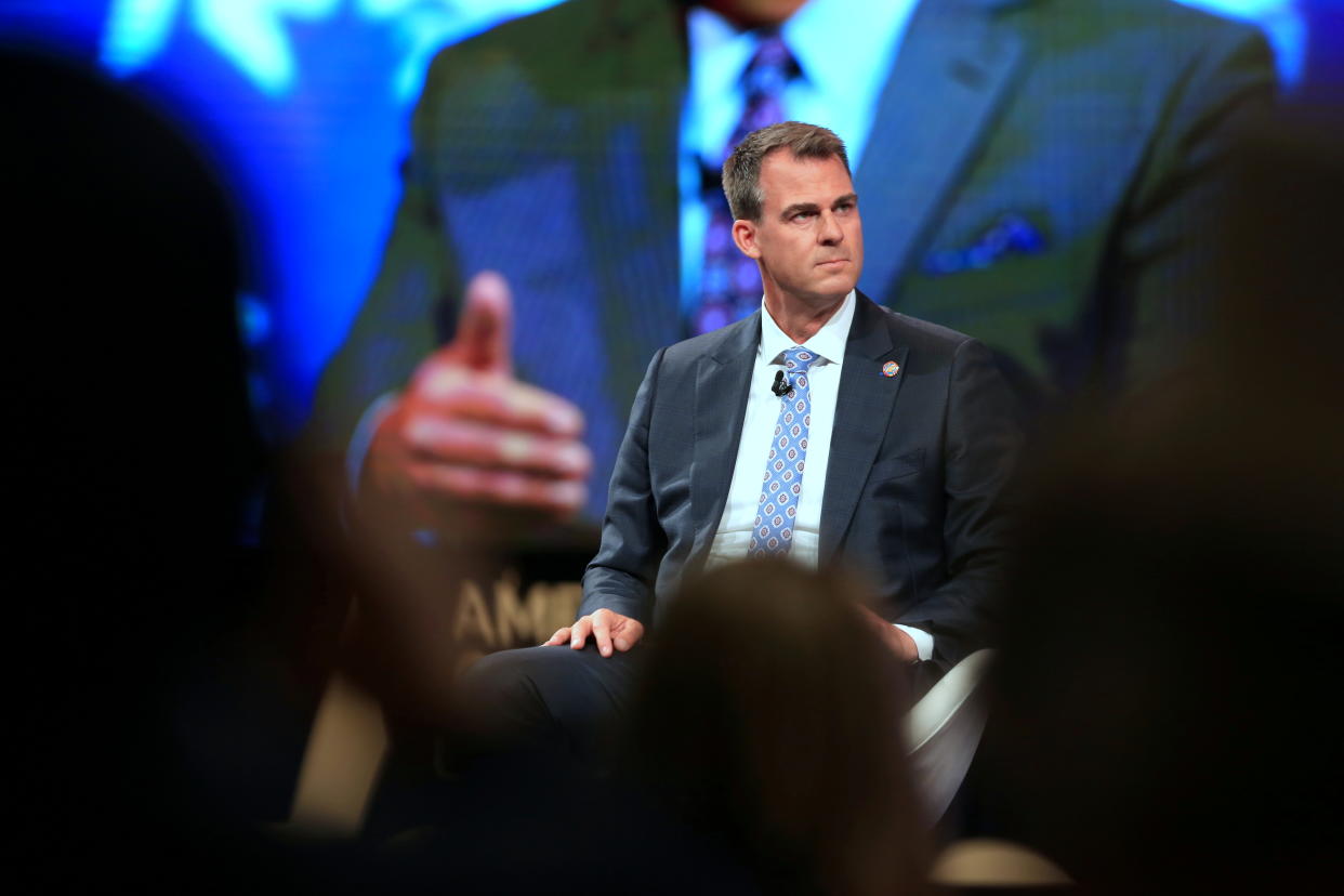 Gov. Kevin Stitt of Oklahoma listens as a CPAC speaker appears on a large screen behind him.