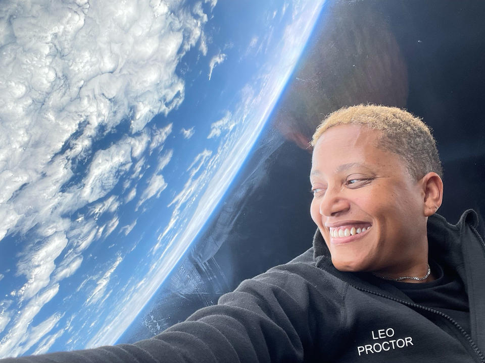 a smiling woman with short hair looks out a spacecraft's window at earth