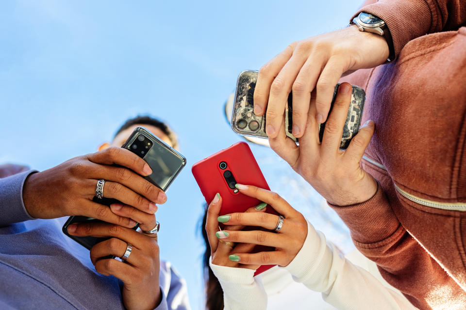Low angle view of three young people using mobile phones outdoors. Addicted gen z friends holding smartphone outside. Blue sky. Copy space for text.