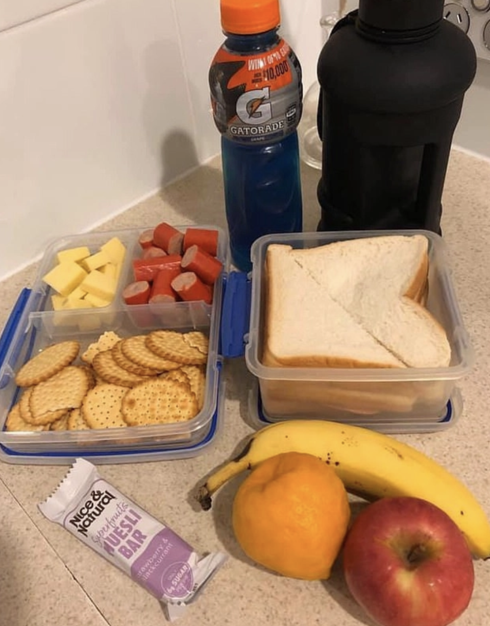 A sandwich in a lunchbox, fruit, Gatorade, sliced cheese and a muesli bar are pictured.