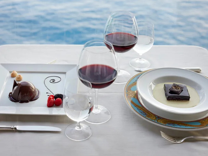 Glasses of wine and water in between two plates of food at a dining table. There are views of the ocean in the back.