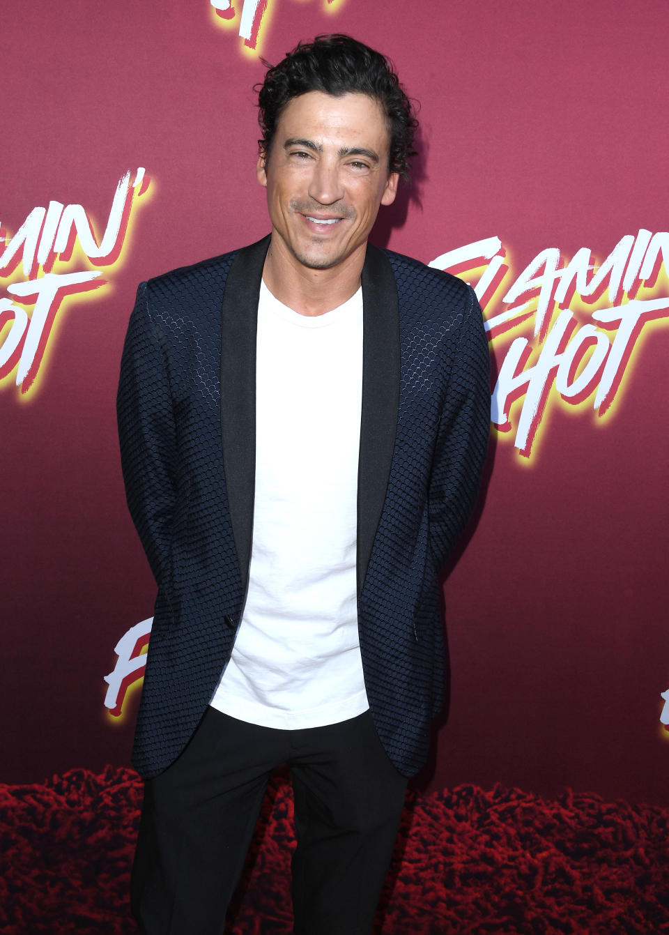 Man in a textured blazer and white shirt poses on a "Flamin' Hot" event backdrop