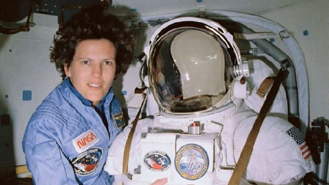 Kathy Sullivan wearing a blue flight suit is positioned next to a spacesuit.