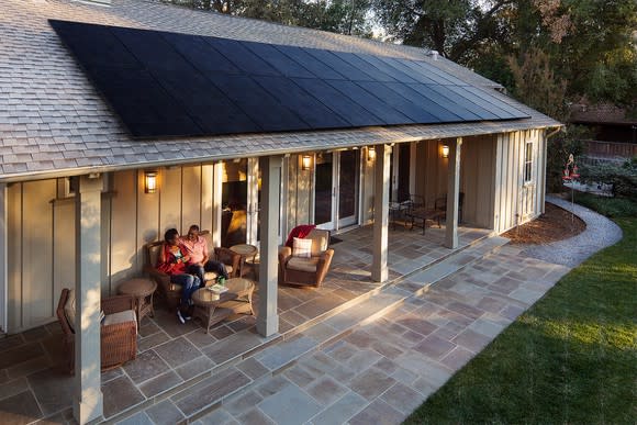 A home with solar panels on the roof and two people sitting under a covered patio.