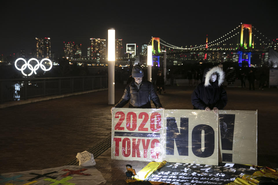 Anti-Olympic activists hold a sign as the illuminated Olympic rings are visible in the distance Friday, Jan. 24, 2020, in the Odaiba district of Tokyo. Tokyo put on a flashy fireworks display on Friday to mark the 6-months-to-go milestone for this summer's Olympics. (AP Photo/Jae C. Hong)