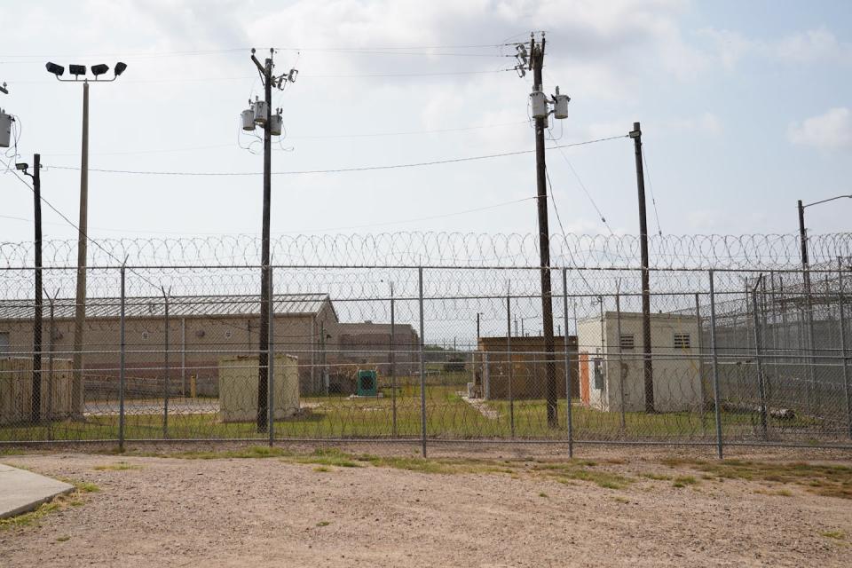 Detainee yard with low buildings behind fences topped with barbed wire and tall light poles