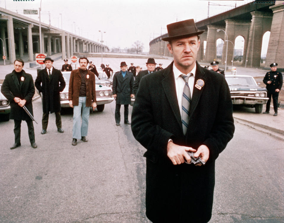 American actor Gene Hackman (foreground), as Detective Jimmy 'Popeye' Doyle, stands in the street by an overpass in front of a group of policemen and holds a gun in his hands in a still from the film 'The French Connection,' directed by William Friedkin, 1971. In the background is actor Roy Scheider (first from left) as Detective Buddy 'Cloudy' Russo and policeman and actor Eddie Egan (1930 - 1995) (fifth from left) as Walt Simonson. (Photo by 20th Century Fox/Hulton Archive/Courtesy of Getty Images) 