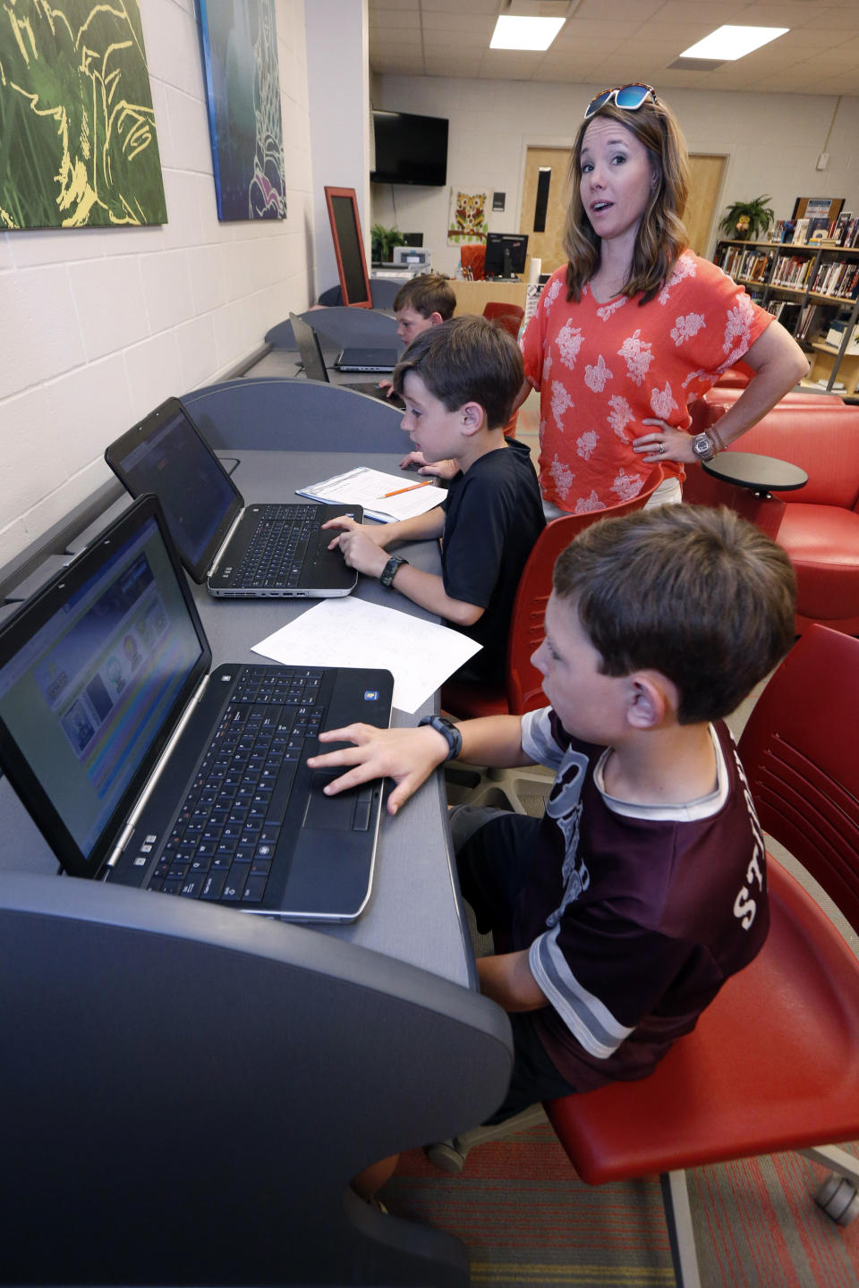 In this May 8, 2019, photograph, Sharon Stidham, standing, talks to her sons Graham, center, and Miles, front, as they use the laptops at East Webster High School to do homework in Maben, Miss. A cellphone tower is visible through the trees from their home on a hilltop near Maben, but the internet signal does not reach their house, despite building a special antenna on top of a nearby family cabin. (AP Photo/Rogelio V. Solis)