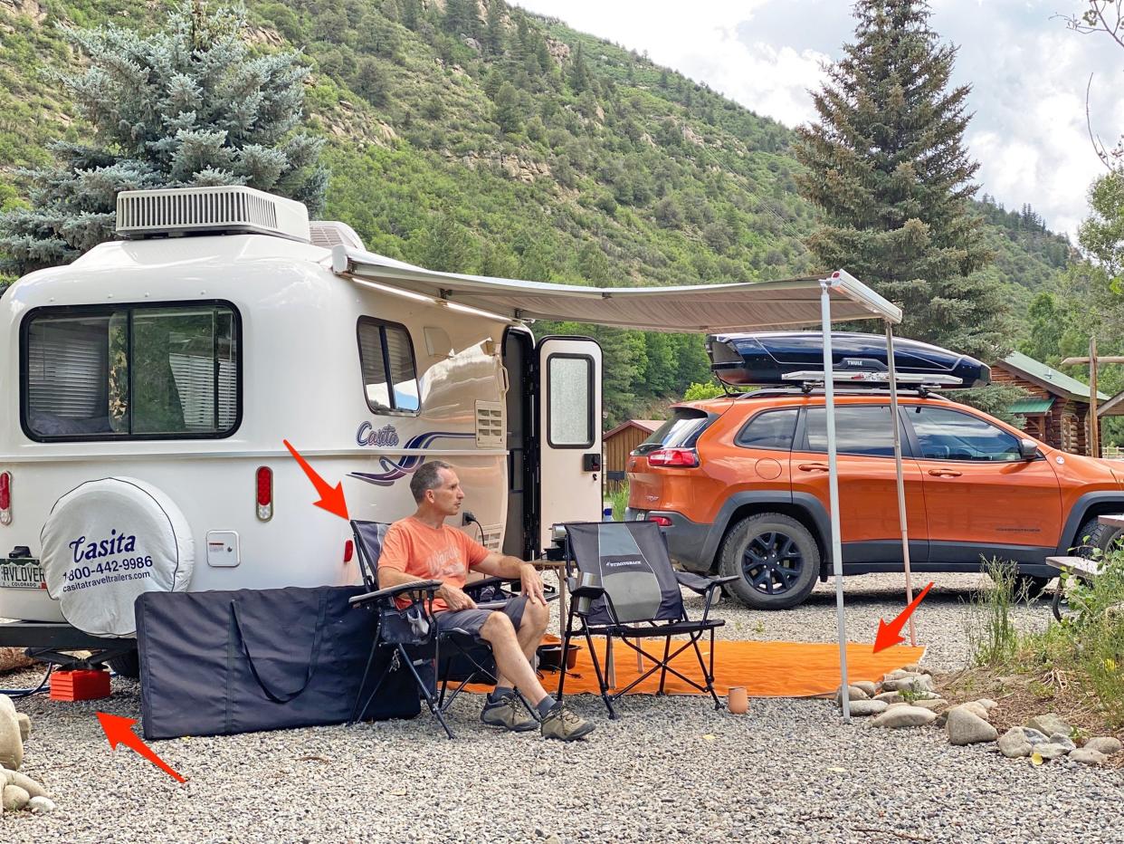 Marc sits at a campsite with his trailer and vehicle. There are mountains and trees behind him. Arrows point to essentials