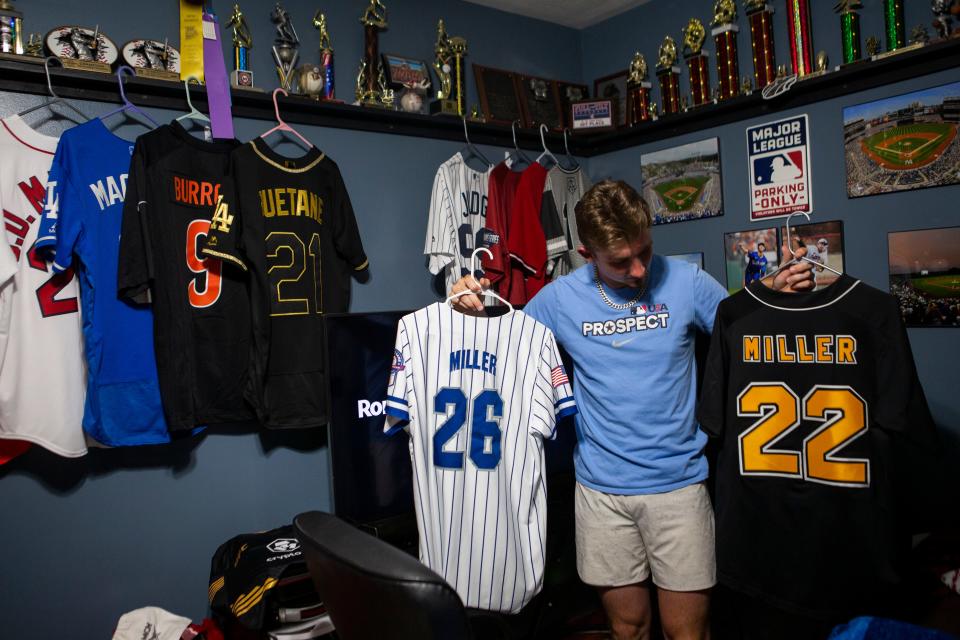 Jacob Miller holds up a couple of the jerseys he has worn  in different national baseball tournaments in his bedroom at his home in Baltimore, Ohio on May 18, 2022. Jacob's room is filled with multiple trophies, jerseys and his personal baseball memrobilia he has collected throughout his career. 