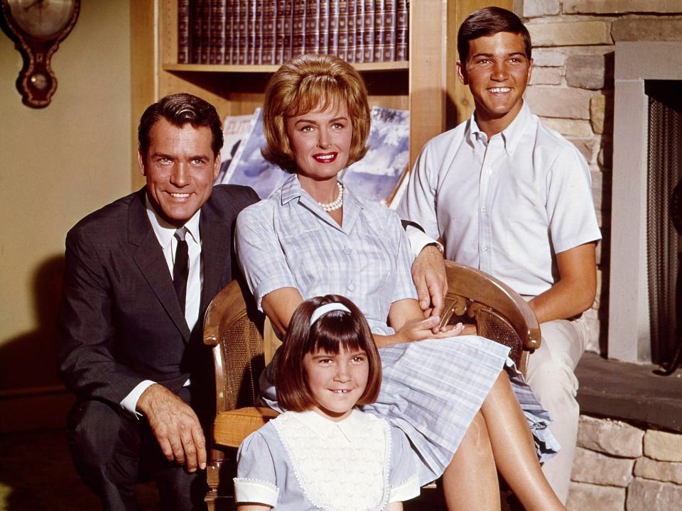 Donna Reed and her TV family sitting on set in a living room with Donna in a chair in the middle of them.