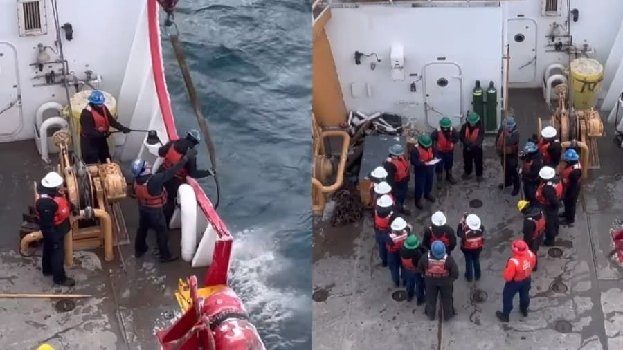 Several crew members use a crane to lower an anchor into the water while the rest of the crew huddle on the deck.