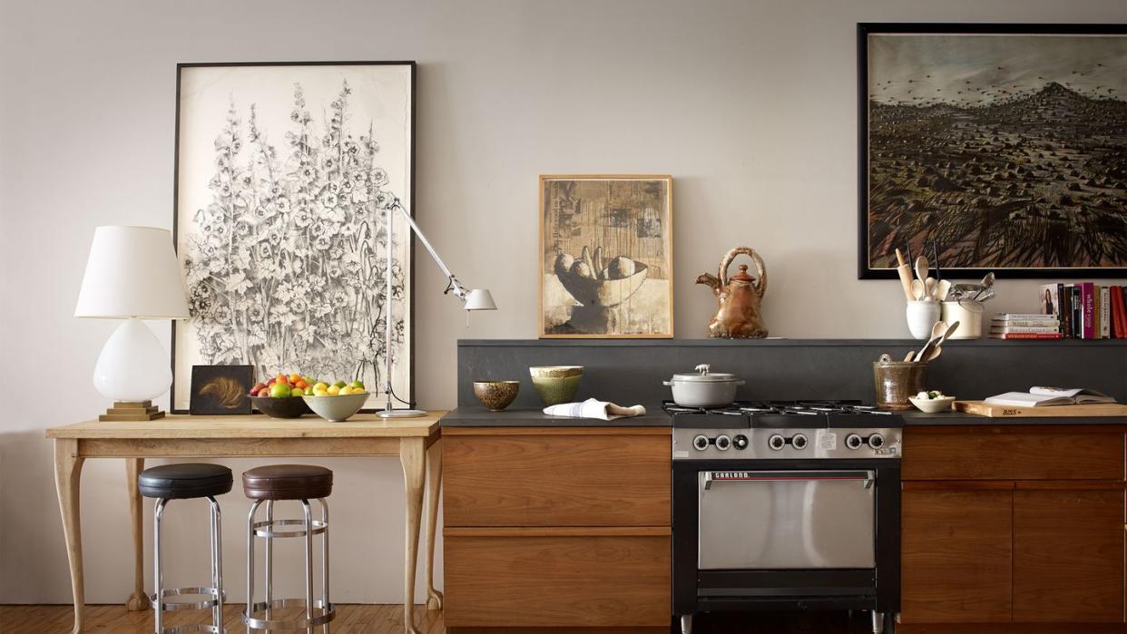 open kitchen with custom cabinetry in a dark honey shade and a small but expensive looking stainless stove and lots of artwork on the wall behind and a long pale colored table adjacent