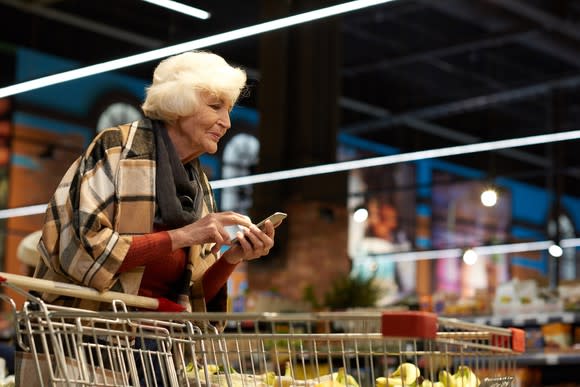 Senior woman typing on phone while pushing shopping cart.