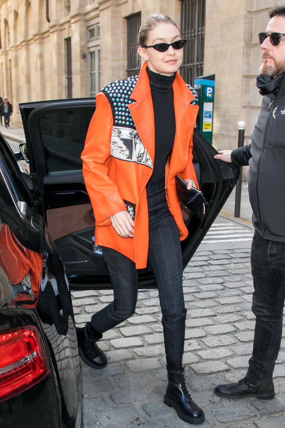 <p>in a bright orange patent leather oversized blazer over a tight black turtleneck, with black plaid pants, lace-up combat boots, and sunglasses.</p>