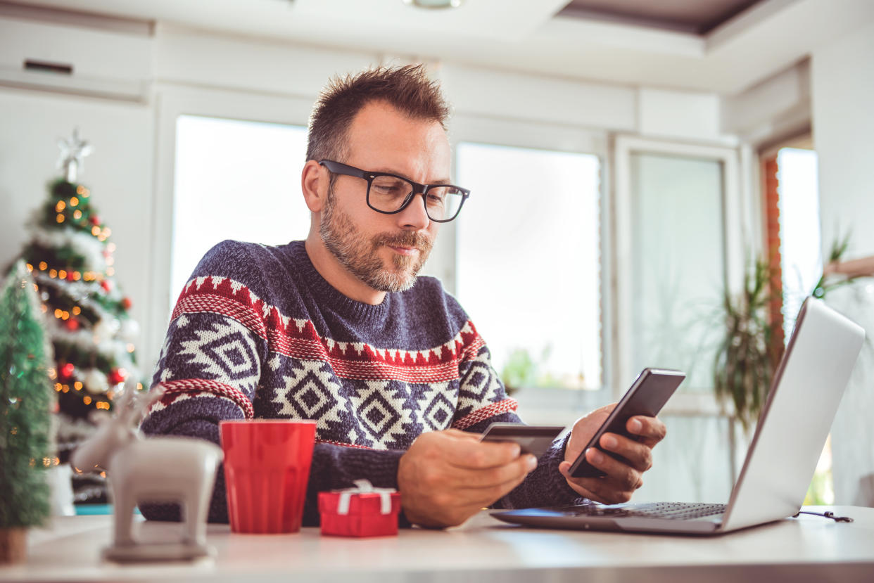 Men wearing blue sweater and eyeglasses holding credit card and using laptop at home office during christmas holidays