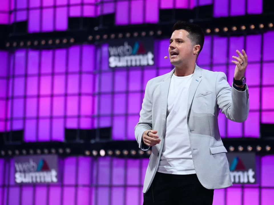 Domm Holland is standing on a stage, the background are purple LED squares with a sign that says "web summit." He's wearing a grey blazer, white tshirt and black pants, his hands are animated as he speaks.