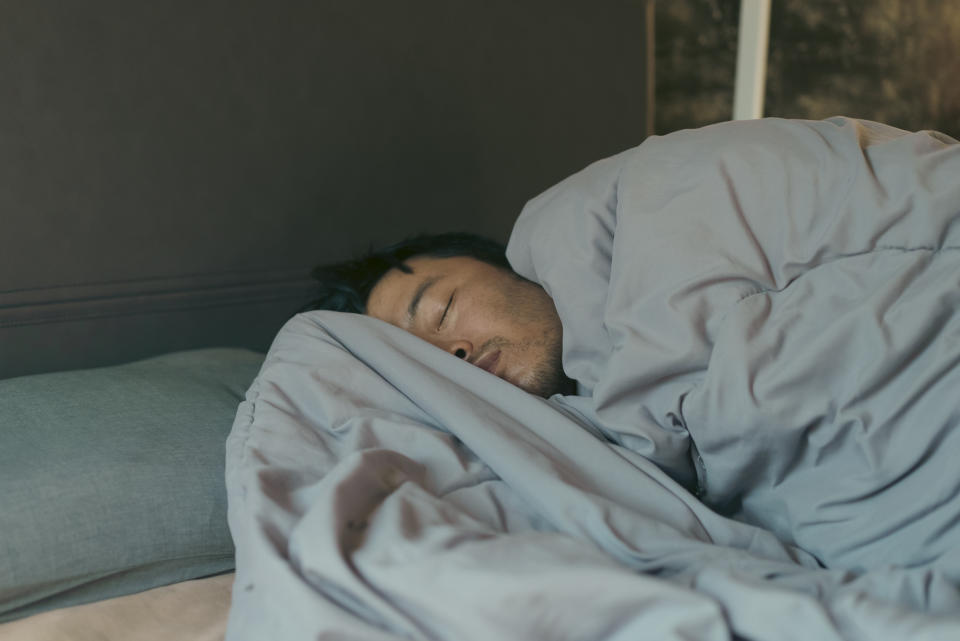 A man is asleep in bed, covered in a blanket. His face is partially visible and he appears relaxed