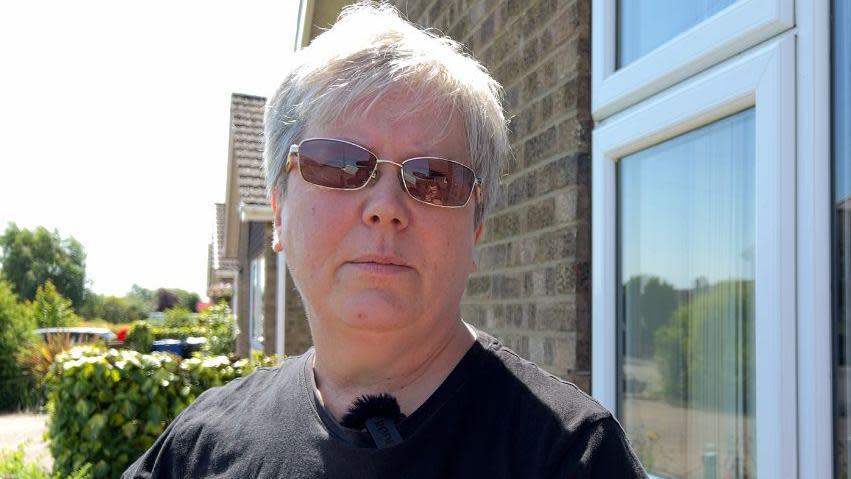 Paula De Vries, with short gray hair, wears sunglasses and a black t-shirt while standing in her front yard