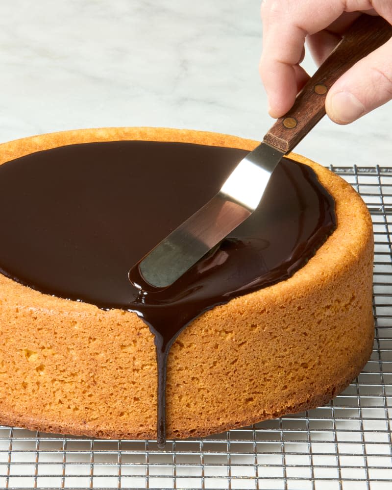 angled shot of a full bolo de cenoura on a cooling rack, with a chocolate glaze being spread on top of the cake.