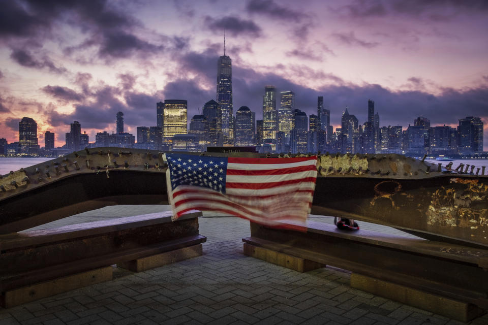 A U.S. flag hanging from a steel girder, damaged in the Sept. 11, 2001 attacks on the World Trade Center, blows in the breeze at a memorial in Jersey City, N.J., Sept. 11, 2019 as the sun rises behind One World Trade Center building and the re-developed area where the Twin Towers of World Trade Center once stood in New York City on the 18th anniversary of the attacks. (AP Photo/J. David Ake)