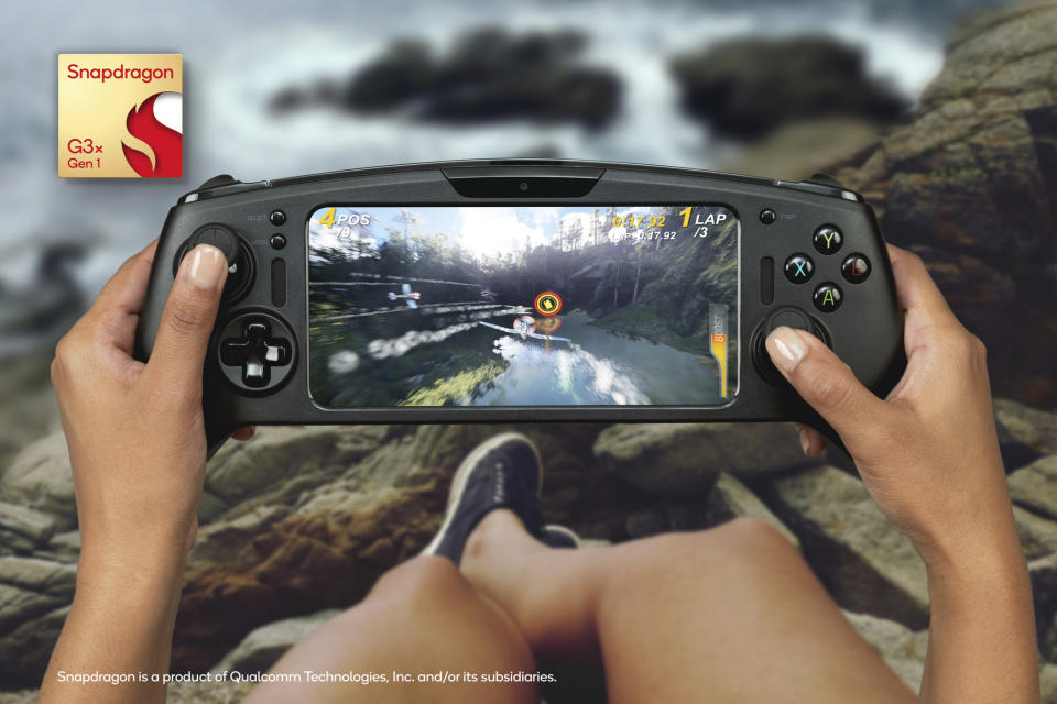 A person sits on rocks by the beach, playing a game on a handheld gaming device created by Razer.