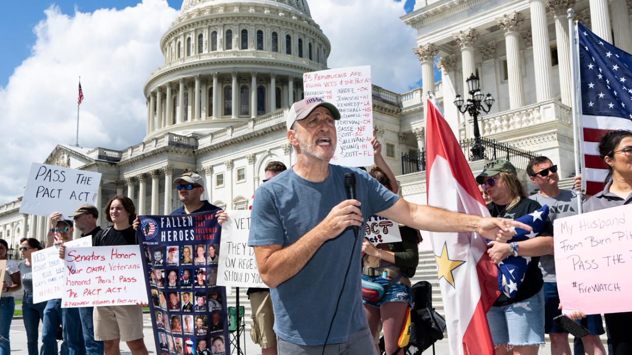 Jon Stewart holds a microphone in front of the U.S. Capitol among protesters holding signs, some of which read: Pass the PACT Act and Senators, honor your oath, veterans honored theirs, and Fallen heroes, the war that followed us home.