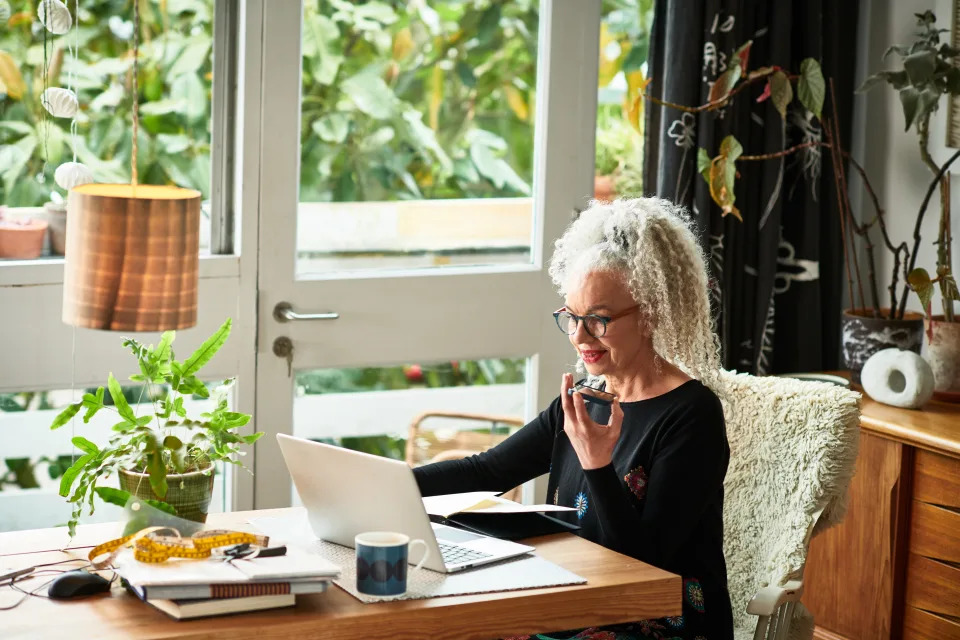 Grey haired female entrepreneur multitasking at home, video conference, speaking and listening on mobile phone, wireless technology