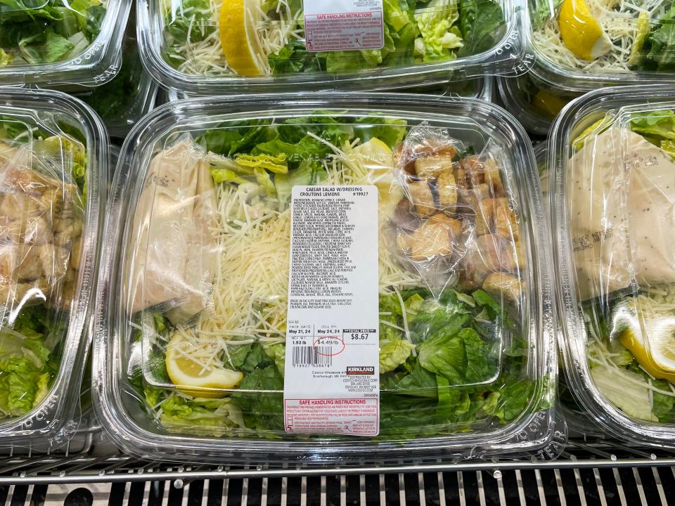 A clear plastic container with lettuce, Parmesan cheese, packets of a white dressing, and packages of croutons