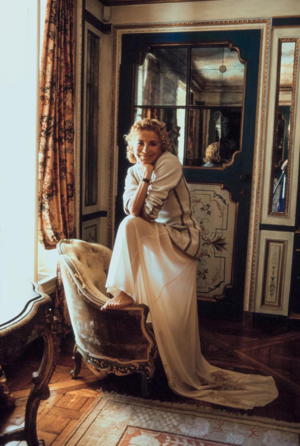 Laura Ungaro, wife of fashion designer Emanuel Ungaro, poses in their Paris, France, home with one foot on up on the arm of a chair and her elbows leaning on her knee. She is wearing a white sweater with a long white skirt. Hair by Kerry Warn at John Frieda using John Frieda products. Makeup by Alan Milroy for Faces.