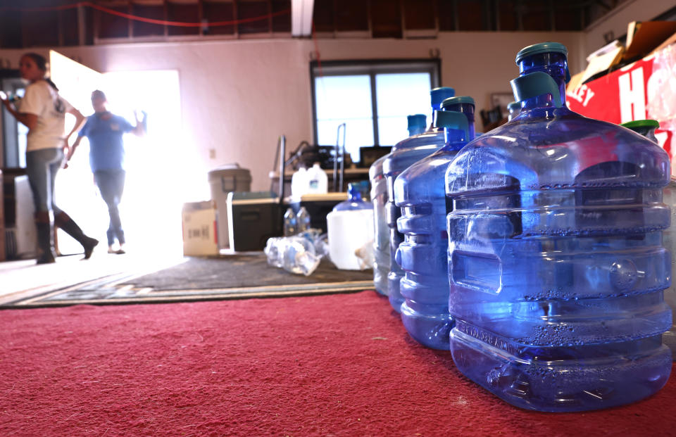 Volunteers stand at an open doorway with sun streaming through it near boxes and water bottles on red carpeting..