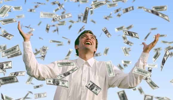Young man smiles and lifts his arms up as money falls down from above.