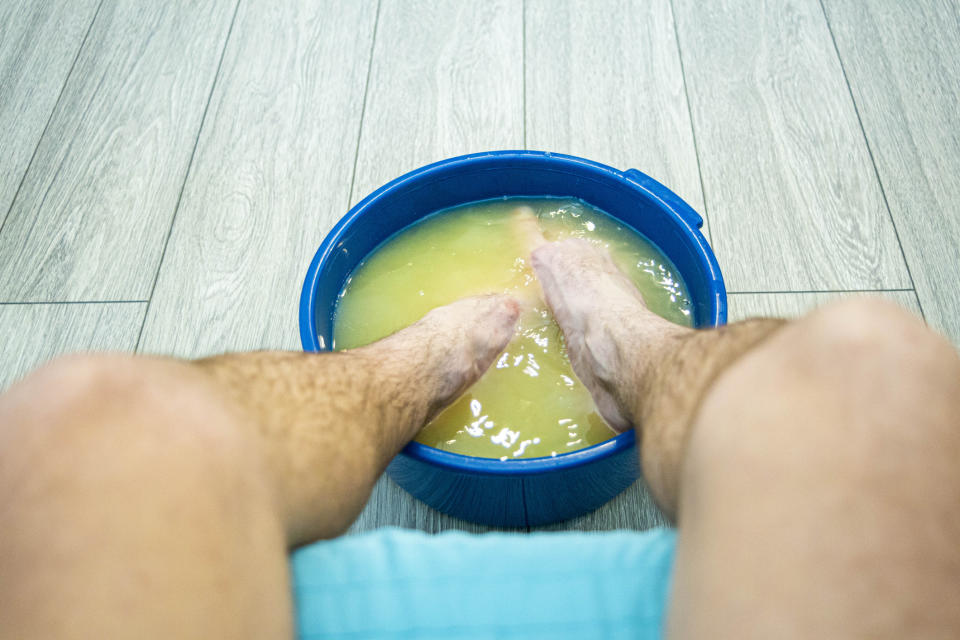 A person soaking their feet in a blue bucket filled with yellowish water on a wooden floor. Only legs and feet are visible