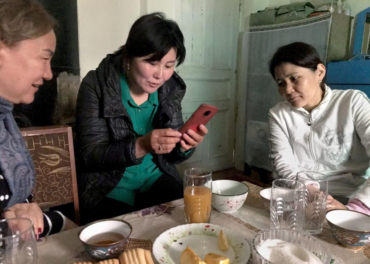 Three women set at a kitchen table, looking at a phone, talking.