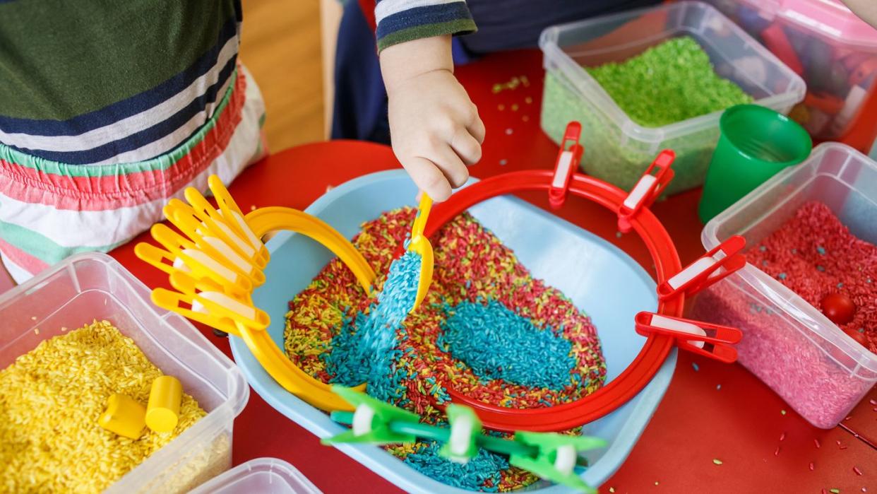 toddlers playing with sensory bin with colourful rice on red table