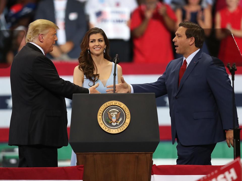 Casey DeSantis behind a lectern as Donald Trump and Ron DeSantis shake hands in front of her.