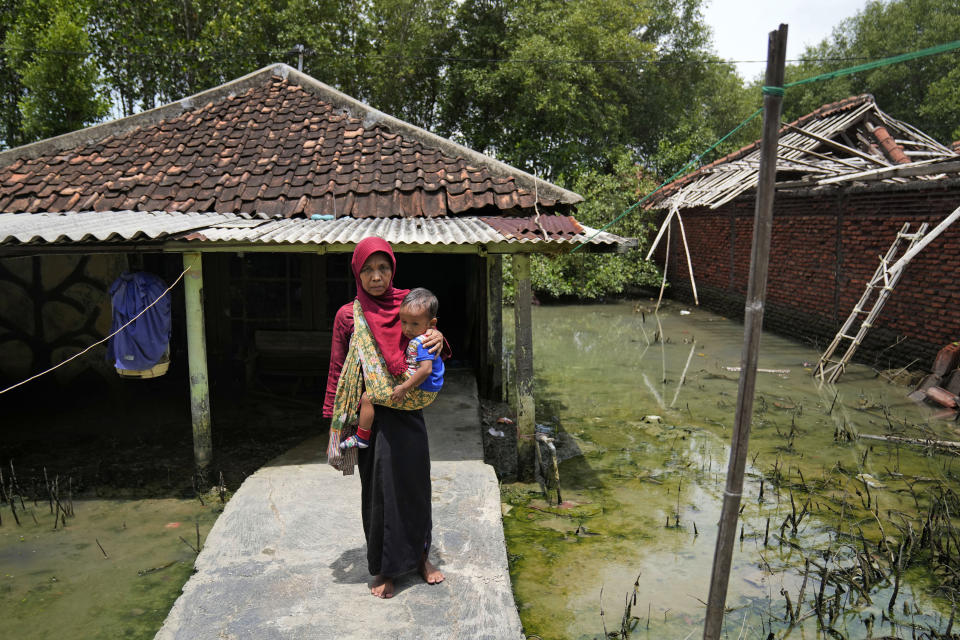 Asturiah, one of the few residents of Mondoliko village who stayed behind after most of her neighbors left due to the rising sea level that inundate their neighborhood on the northern coast of Java Island, carries her grandson Muhammad Rizki Aditya, in front of her house in Mondoliko, Central Java, Indonesia, Monday, Nov. 8, 2021. World leaders are gathered in Scotland at a United Nations climate summit, known as COP26, to push nations to ratchet up their efforts to curb climate change. Experts say the amount of energy unleashed by planetary warming would melt much of the planet's ice, raise global sea levels and greatly increase the likelihood and extreme weather events. (AP Photo/Dita Alangkara)