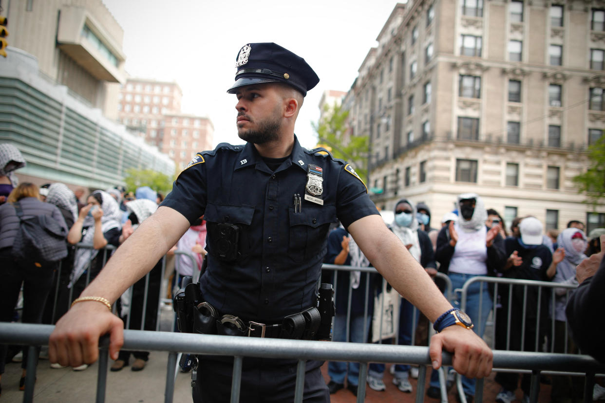 A police officer stands at a temporary fence with a crowd to his back.