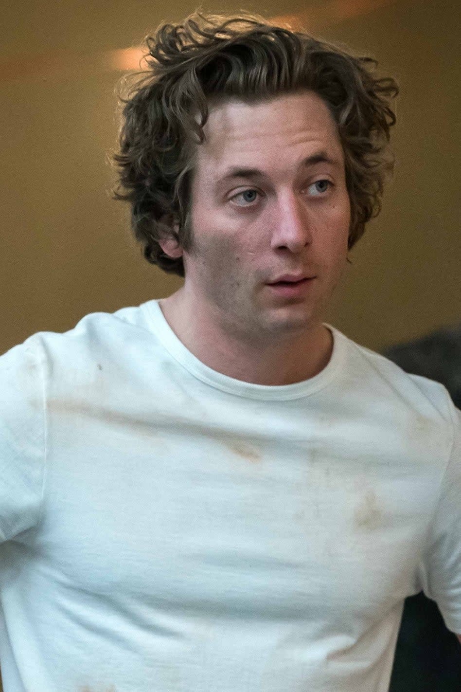 Jeremy Allen White stands with hands on hips in a casual, work-stained white shirt. A man in a black shirt and beanie stands blurred in the background