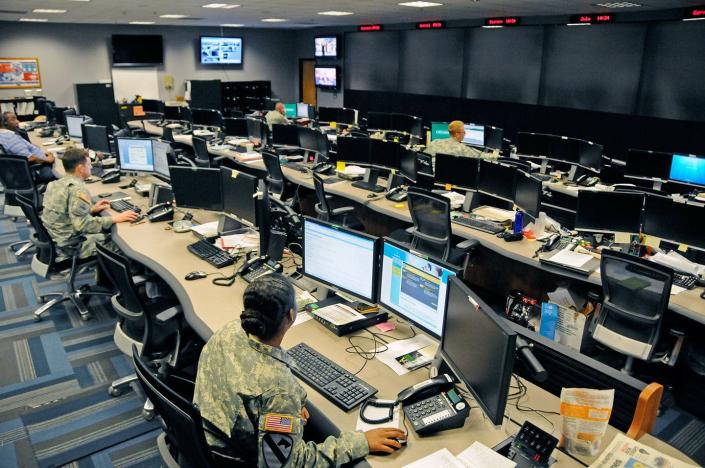 people in military uniforms sit at desks with multiple computer monitors