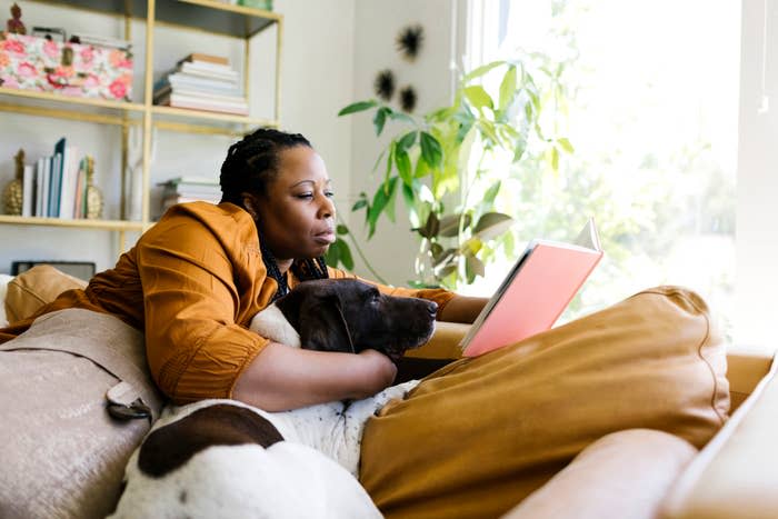 A person lying on a couch, reading a book with a dog resting beside them. Shelves with books and plants are in the background