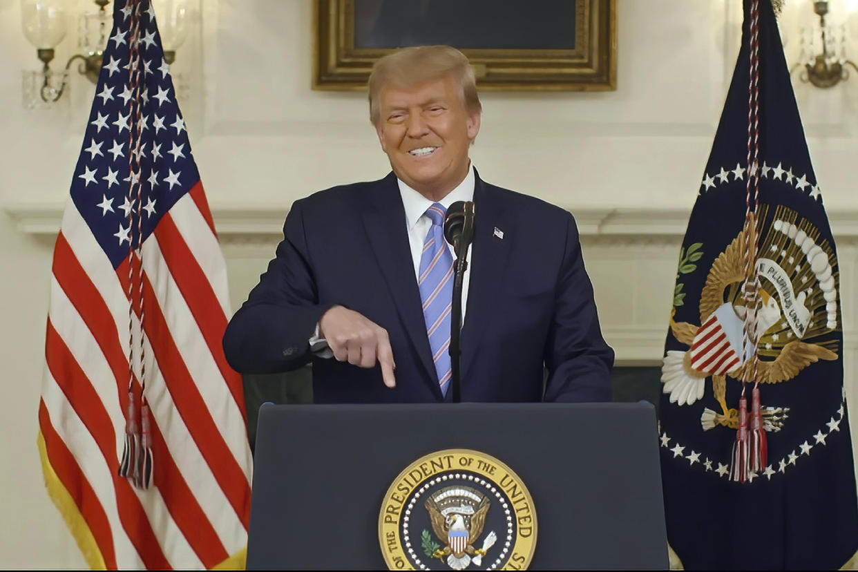 Donald Trump stands at a podium with the Presidential Seal and a microphone and points a finger downward as he makes a pained grimace of apparent frustration.
