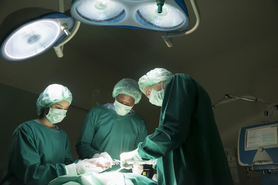 Three surgeons in green surgical gowns and caps are performing an operation under bright surgical lights in an operating room