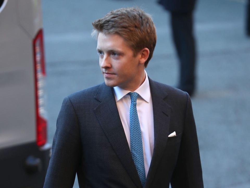 Hugh Grosvenor walking behind a grey van while wearing a dark blue suit and a light blue tie.